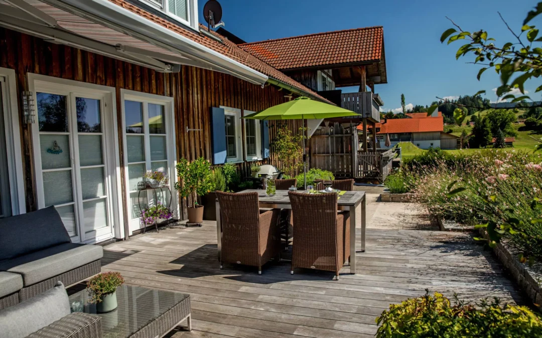 Holzterrasse mit einem Tisch, Stühlen und einem grünen Sonnenschirm in einem Gartenbereich.