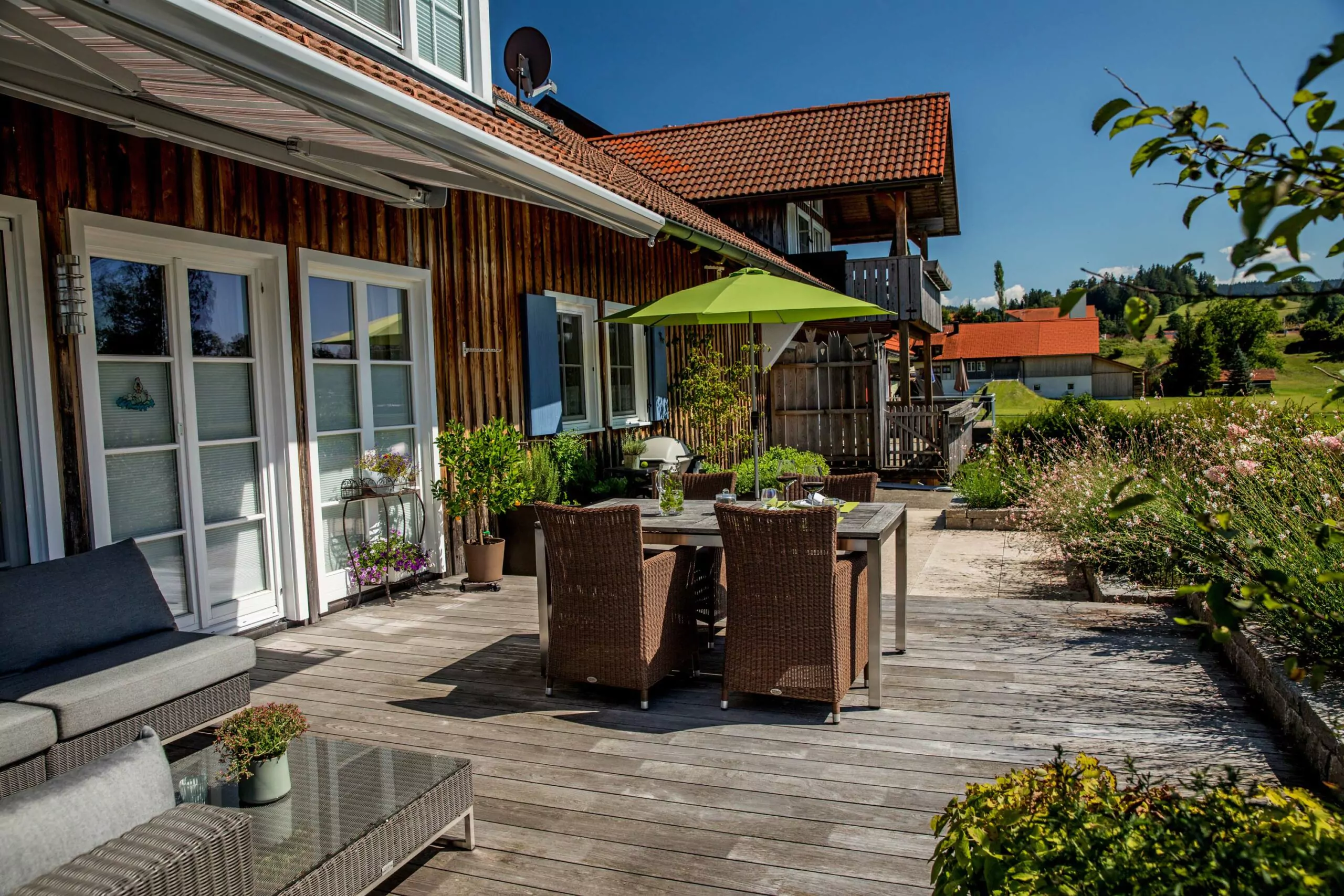 Holzterrasse mit einem Tisch, Stühlen und einem grünen Sonnenschirm in einem Gartenbereich.