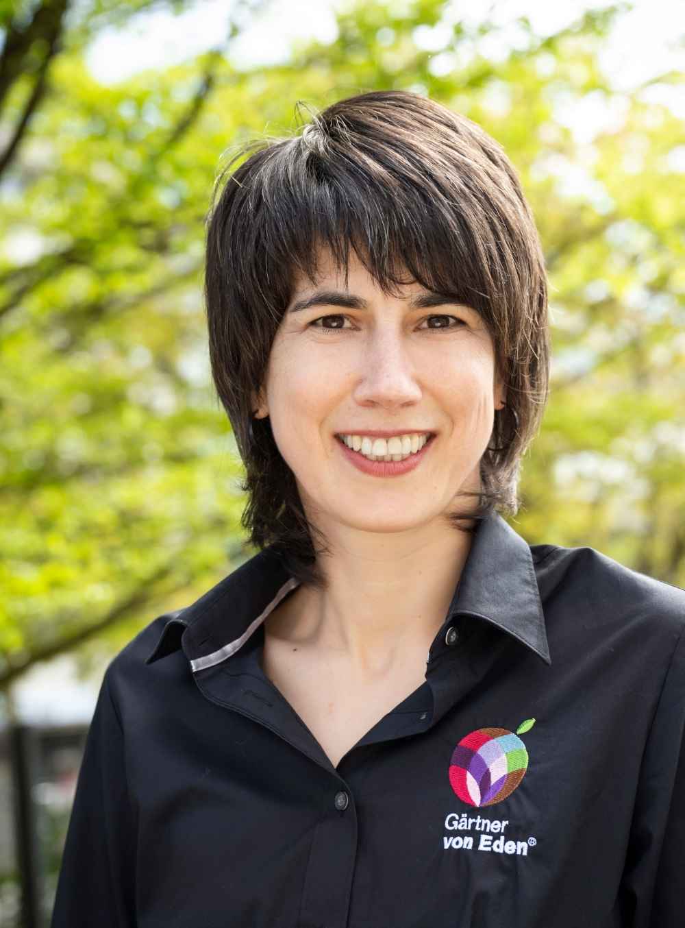 Sonja Hoeps lächelt im Freien vor grünen Bäumen und trägt ein schwarzes Hemd mit dem Logo von Gärtner von Eden.